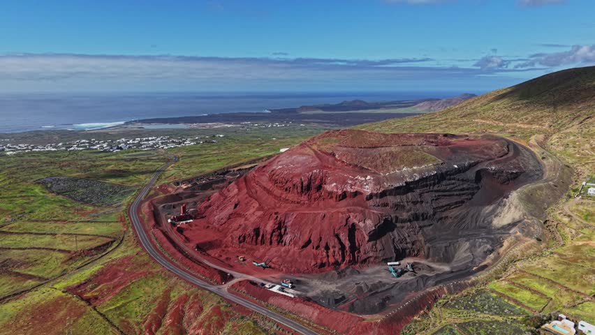 A large volcanic hill with red soil stands out against the blue sky and ocean in Lanzarote, Spain. Vehicles can be seen working near the hill in this active area.