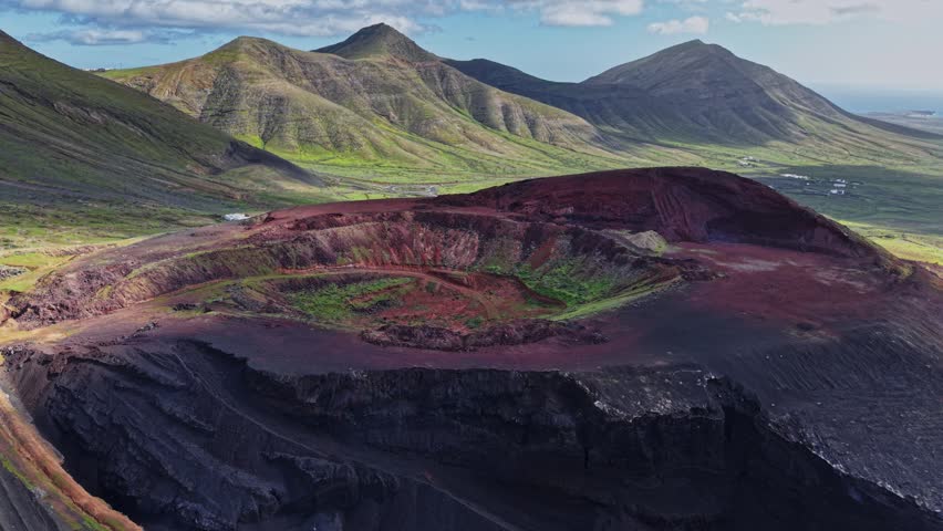 A view of a large volcanic crater in Lanzarote shows the red and black earth. Nearby hills create a scenic backdrop under a clear sky in daytime. Green vegetation grows in the crater.