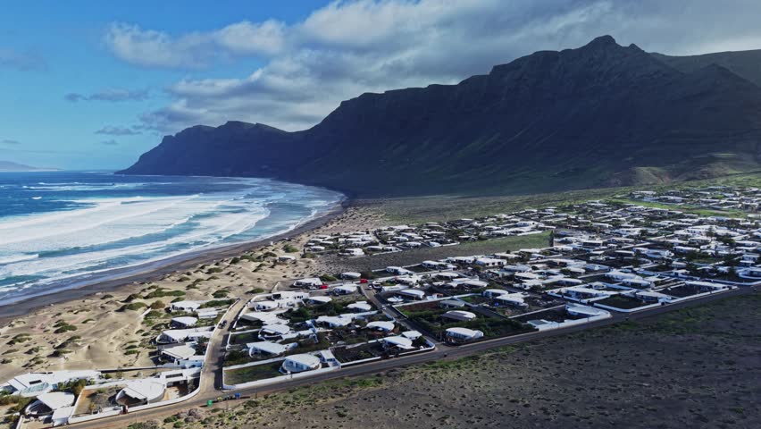 A coastal area in Lanzarote, Spain. The beach stretches along the shore with waves breaking. Houses are built close to the sand with hills in the background.