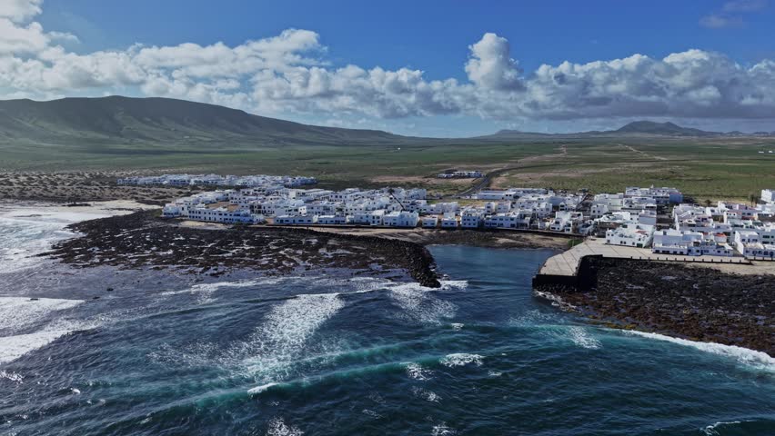 The coastal town sits near the ocean with white buildings lining the shore. Waves crash onto rocks while a dock extends into the water. Mountains rise in the background with clouds overhead.