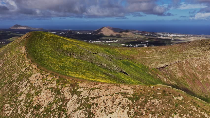 View of layered mountains and green fields near the coast of Lanzarote. White houses sit among the fields under a bright blue sky with some clouds. It is midday.