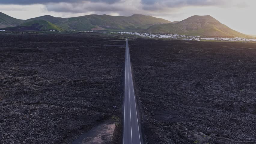 A road stretches through a volcanic landscape in Lanzarote, Spain. Fields and mountains surround the path. A small town is visible in the distance under cloudy skies around sunset.
