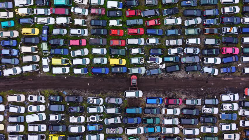 Cars are parked closely together in a junkyard with a dirt path running through the middle. Different colors and makes of vehicles fill the area under bright sunlight.