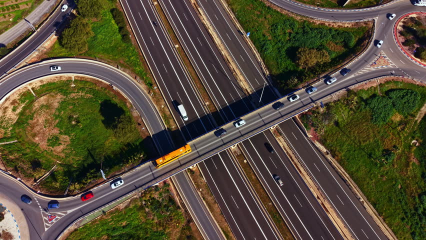 Cars move along highways and bridges in an urban area. The layout shows multiple lanes and an intersection where roads connect. Green spaces are nearby.