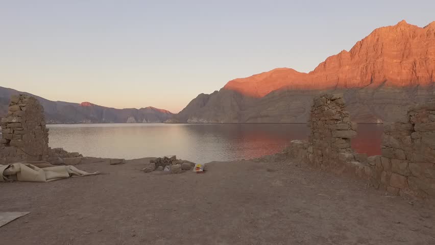 Drone advances slowly from historic Telegraph Island ruins and campfire to calm water at sunset in Strait of Hormuz between Iran and Oman, revealing peaceful remote landscape