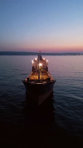 Cinematic drone shot of an oil tanker ship anchored at sea during twilight. The vessel is illuminated by onboard lights with a beautiful city skyline and sunset glow in the background.