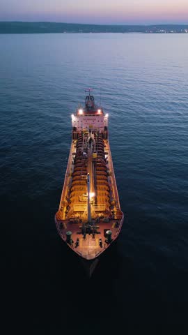 Cinematic drone shot of an oil tanker ship anchored at sea during twilight. The vessel is illuminated by onboard lights with a beautiful city skyline and sunset glow in the background.