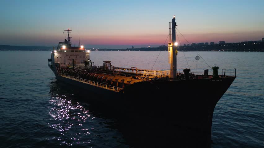 Cinematic drone shot of an oil tanker ship anchored at sea during twilight. The vessel is illuminated by onboard lights with a beautiful city skyline and sunset glow in the background.