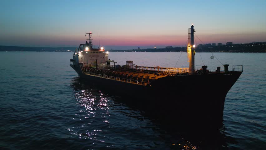 Cinematic drone shot of an oil tanker ship anchored at sea during twilight. The vessel is illuminated by onboard lights with a beautiful city skyline and sunset glow in the background.