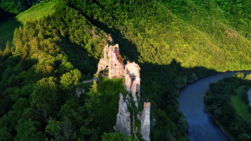 Approaching the ruins of the ancient castle lit by the bright sun. Amazing wooded Tatra mountains and narrow river at backdrop. Aerial view.