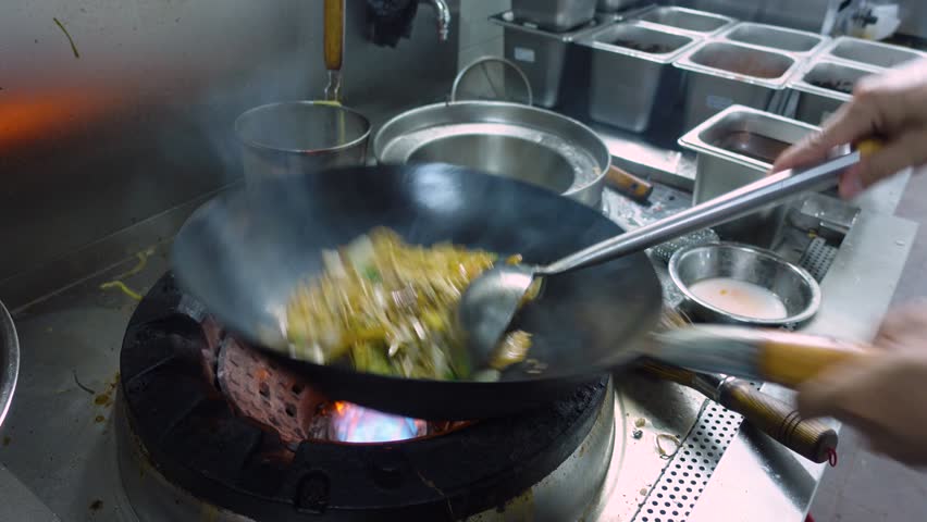 Chef Adding Seasoning While Tossing Noodles And Vegetables In Wok Over Flame At Restaurant Kitchen. close-up shot