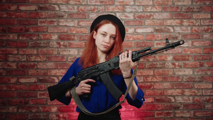 A portrait of a confident caucasian young redhead woman in a blue blouse and black beret, holding a black Kalashnikov assault rifle and looking at the camera with a fighting spirit against brick wall