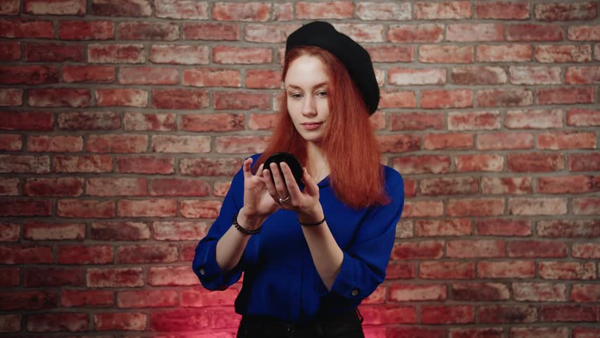 A portrait of an attractive caucasian young redhead woman in a blue blouse and black beret, preens and applies powder to her face while looking in pocket mirror against brick wall. Maintaining beauty