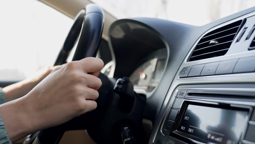 Woman's hands gripping the steering wheel while driving a modern car on a sunny day, with the dashboard, instrument panel, and air conditioning vents visible in the foreground
