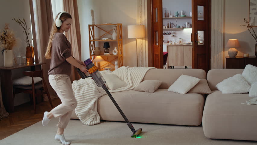 Woman vacuums carpet in living room while wearing headphones. Young housewife sways with music as guides cordless cleaner near sofa with playful energy