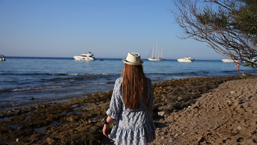 Back view of Young Woman in Summer Dress and Straw Hat Looking at Sea. Romantic Vacation Concept, Tropical Beach Lifestyle, Female Traveler enjoying Ocean View with Boats, Freedom,Serenity,Wanderlust