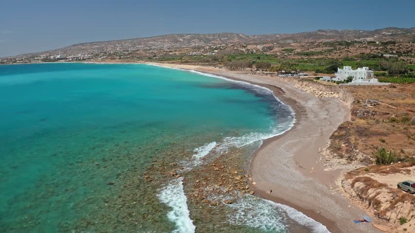 Cyprus coastline with beautiful turquoise Mediterranean sea meeting a long pebble beach, with a white building on the rocky shore under a clear summer sky during a sunny day. Aerial view drone flight