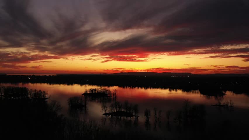 Drone crane shot moving down slowly behind the black silhouette of bare trees, at the end of sunset, with dramatic colors of orange, gold, and turquoise contrasting with the black trees in the calm river