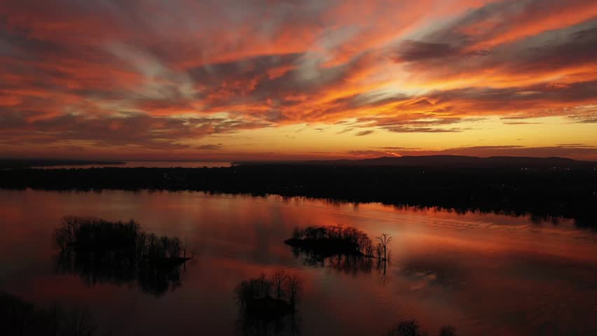 High altitude drone shot, gently receding revealing the splendid sunset, with dramatic colors of orange, gold and contrasting with the black trees in the calm river