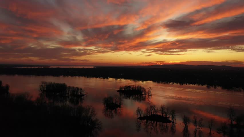 High altitude drone shot, moving slowly forward to show the stunning sunset, with dramatic colors of orange, gold, and contrasting with the black trees in the calm river