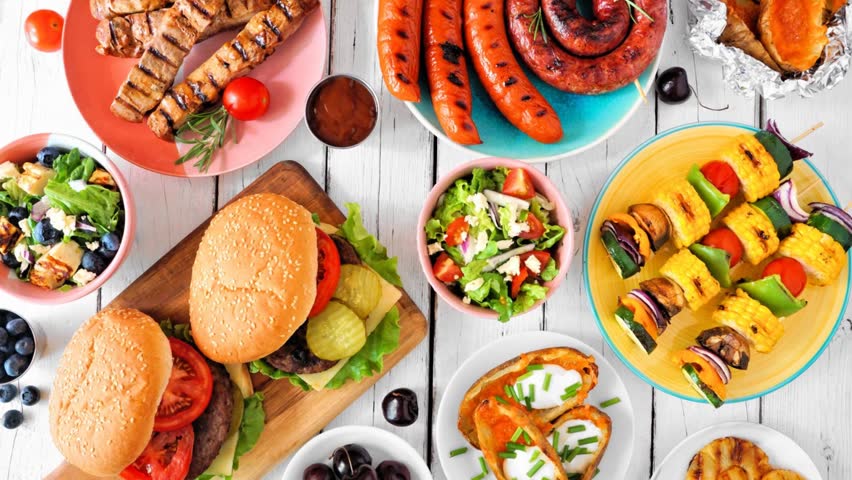 Summer BBQ or picnic food table scene. Slow rotating zoom motion. Assortment of burgers, grilled meat, vegetables, fruits, salad and potatoes. Overhead view on a white wood background.