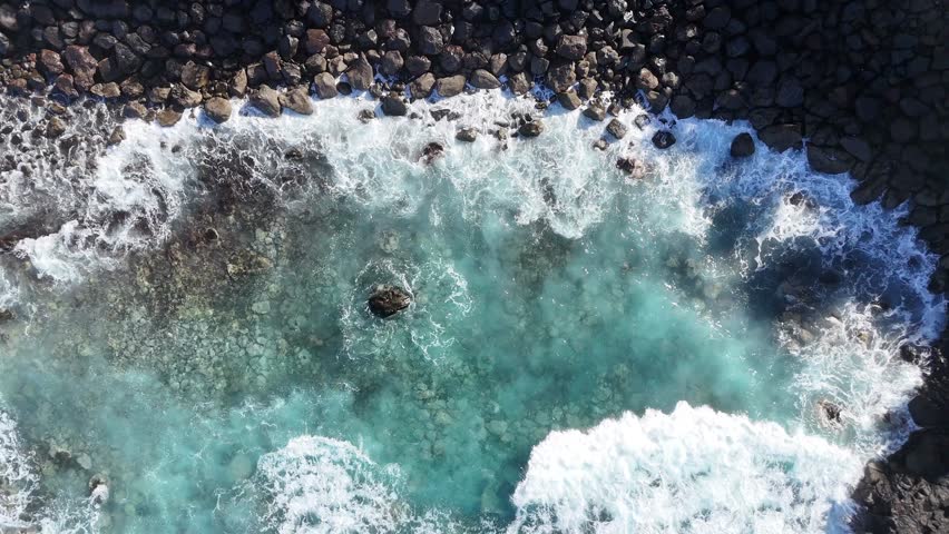 Aerial view of turquoise ocean waves crashing against rocky shoreline. Dynamic movement of water and foam in coastal environment with clear blue hues. Vibrant turquoise water in a coastal setting