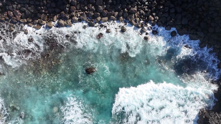 Aerial view of turquoise ocean waves crashing against rocky shoreline. Dynamic movement of water and foam in coastal environment with clear blue hues. Vibrant turquoise water in a coastal setting