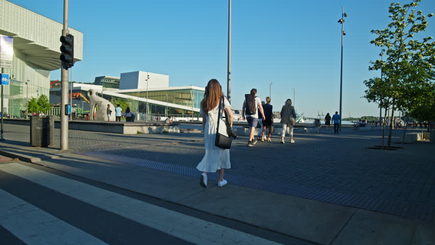 Tourist woman walking toward Oslo Opera House across open plaza in Oslo. Modern architecture and waterfront area during summer in Norway.