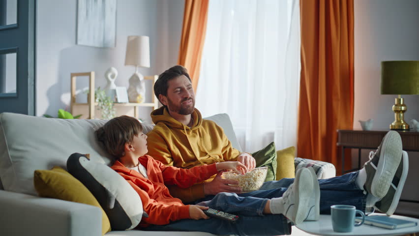Smiling father playing popcorn watching television movie together at relaxed family leisure closeup. Carefree playful boy eating snacks enjoy TV viewing relaxing home couch. Parent child domestic life