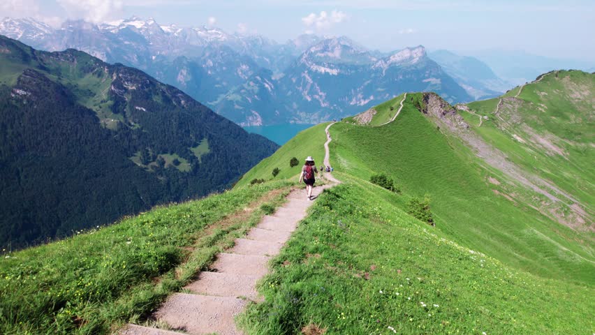 Aerial 4K drone view of hikers walking along the grassy Stoos Ridge in Switzerland, with sweeping alpine mountains, deep valleys, and clear summer skies creating a stunning scenic panorama.