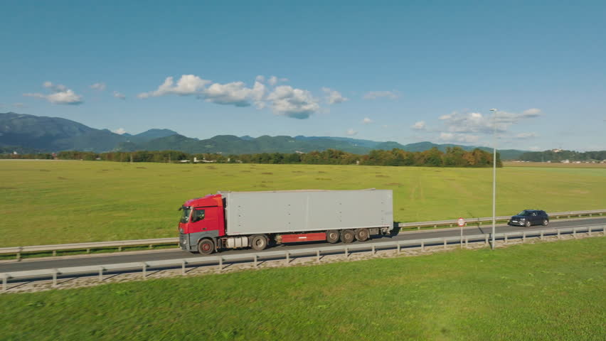 A red truck drives on a highway next to wide green fields and mountains under a clear sky. Another car also travels the same road.