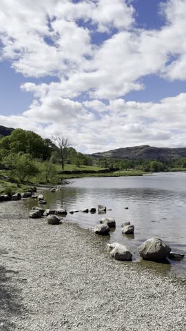 Shoreline footage of Rydal Water with scattered rocks, calm water and surrounding hills under a partly cloudy sky, Lake District, England, UK.