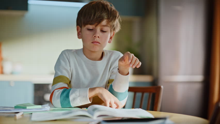 Sad pupil tired homeschooling sitting home desk closeup. Sleepy schoolboy learning homework reading textbook in apartment kitchen. Fatigued little boy unmotivated domestic studying feeling boredom.
