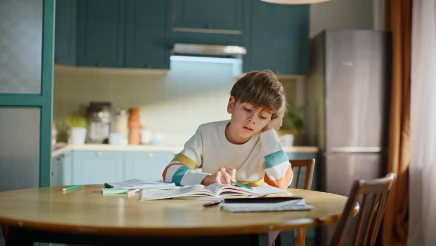 Fatigued schoolboy reading textbook sitting kitchen desk. Tired primary school pupil looking book overwhelmed lesson assignment home. Bored little boy studying alone unmotivated to learning routine.