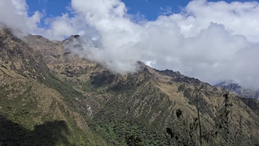 Cusco Region, Peru - 26 June 2024. Clouds hang over steep Andes mountains along the Inca Trail, with rocky ridges, patches of vegetation, and shifting shadows under a sky.