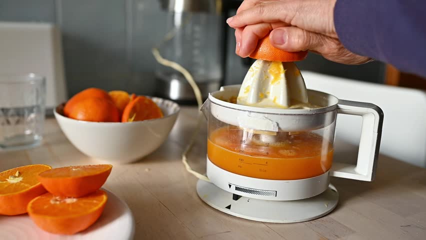 Close-up from a different angle of a woman preparing fresh orange juice with an electric juicer in a home kitchen. Concept of vitamin C intake, immune system support and natural health habits