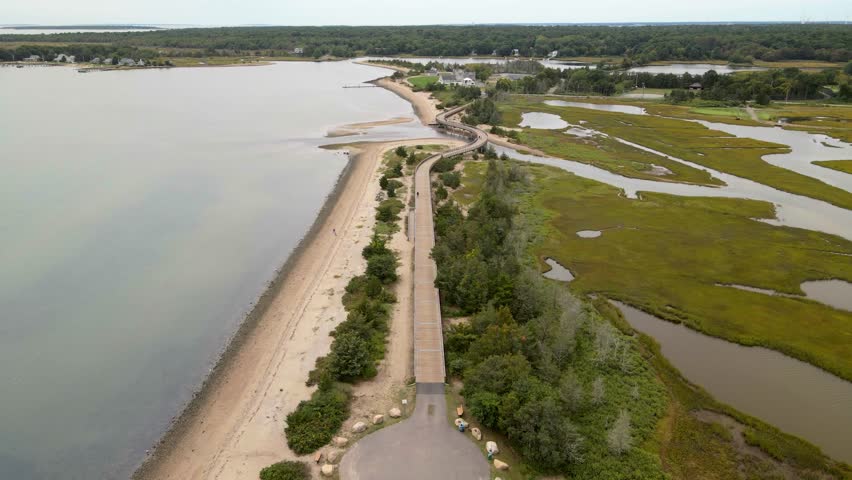 Drone footage captures a winding wooden boardwalk stretching along a sandy beach and through lush green salt marshes with calm waters.