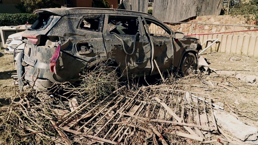 A completely smashed and destroyed car, with broken windows and splashes of mud, standing in a parking lot next to a heavy missile falling site, during the Israel Iran war. Arad, Israel.