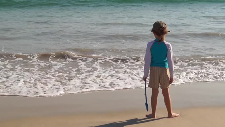 A young girl stands with her back to the camera, looking out at the turquoise sea and white waves, holding a blue shovel on a sunny beach.
