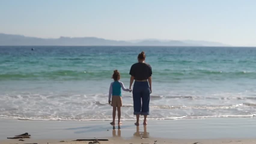 A rear view of a mother and her young daughter standing hand-in-hand on a sandy beach, peacefully watching the turquoise ocean waves on a bright sunny day.