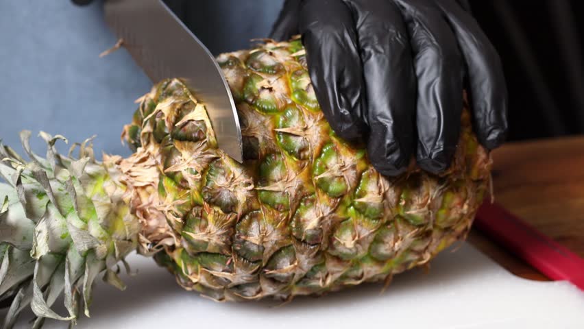 A chef wearing black gloves cuts pineapple with a knife on a cutting board.