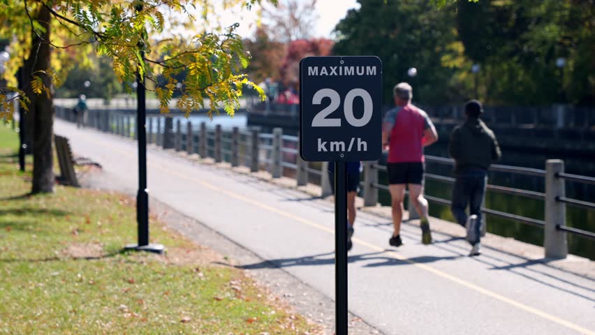 Maximum 20 km h speed limit sign on pedestrian walkway. Joggers running on trail in Ottawa along Rideau Canal in autumn.