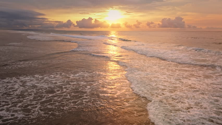Golden sunset sun over Green Bowl Beach, Bali. Warm light reflecting on the Indian Ocean, foamy waves roll toward shore beneath a dramatic bright colorful sky. Natural background. Aerial drone flight