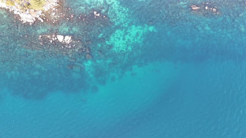 Dramatic aerial top-down view of a scenic highway winding along the rugged shoreline of Lake Tahoe, featuring crystal clear turquoise waters, massive granite boulders, and a lush pine forest.