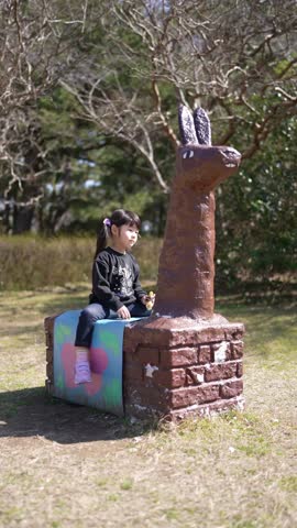 Little girl sits beside whimsical statue. Statue stands tall, crafted from earthy brown material. Base features colorful cube, painted with soft pastel hues. Child smiles gently, gazing toward camera