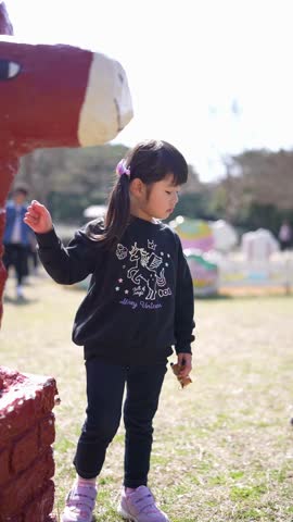 Young girl stands near a giant red sculpture. She gazes upward, curious and attentive. Her pigtails swing slightly with her movement. Soft sunlight illuminates the grassy park. Background figures walk