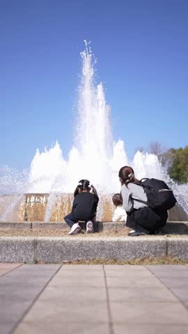 Two children crouch by a powerful fountain. Woman sits nearby, watching them. Clear blue sky frames the water spray. They enjoy the splash, the sun, the sound