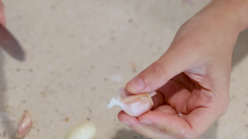Detailed close-up of hands using a knife to peel garlic cloves on a light countertop.