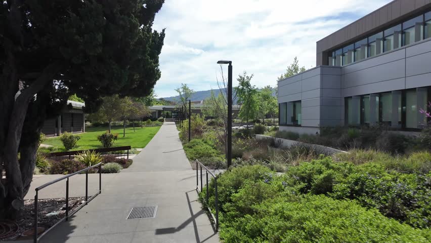 First-person perspective walking along a paved path through the green lawns and modern buildings of the West Valley College campus in Saratoga, California on a sunny day