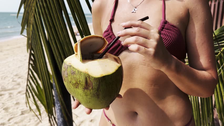 Close up of woman drinking coconut water on beach with palm trees. Tropical holiday, refreshing natural drink and summer lifestyle with sea, palm trees and sunny outdoor vibes